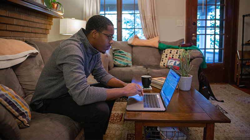 Man in a living room on the sofa looking at a laptop that sits on a coffee table