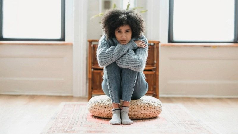 A person sitting cross-legged on a round cushion in a room with two windows. They are wearing a gray sweater, blue jeans, and gray socks.