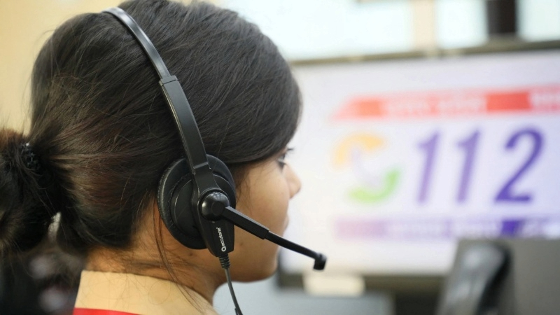 A person wearing a headset, possibly working in a call center. A screen in the background displays the number '112' prominently.