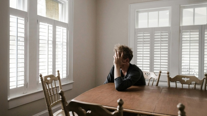 Person sitting at a wooden dining table with their head in one hand in a bright room with white walls and large shuttered windows.
