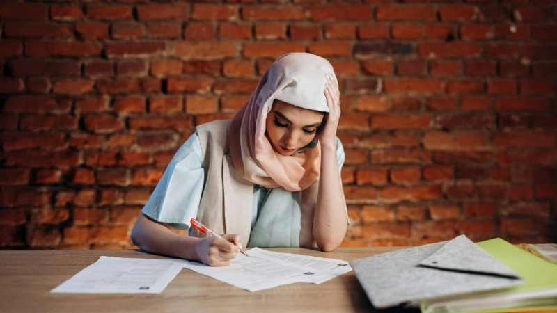 Person sitting at a table with papers, holding a red pen, in front of a brick wall.