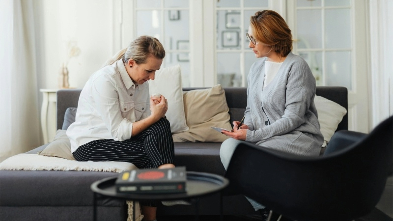 Two people sitting on a couch, one holding a clipboard, possibly in a counseling session. A small table with books is in the foreground.