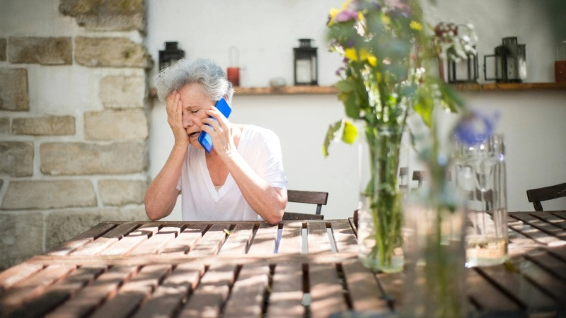 Person with gray hair sitting at an outdoor table, holding a blue phone to their ear. Behind them is a white wall with decorative lanterns on a wooden shelf and a vase of flowers to the right.