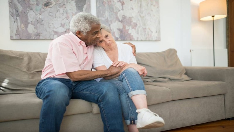 Two elderly people on a beige couch, one with an arm around the other. Abstract art and a floor lamp in the background.