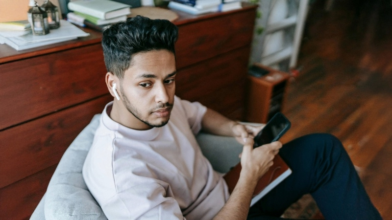 Person sitting on a chair with a smartphone and wireless earbuds. Books and items are on a wooden surface behind them.