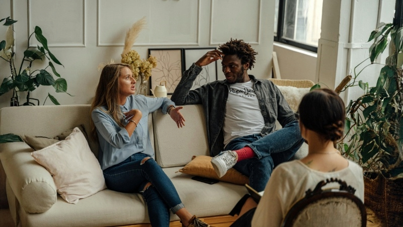 Three people in a living room, two on a beige couch and one facing them on a chair. The room includes plants, framed artwork, and cushions.