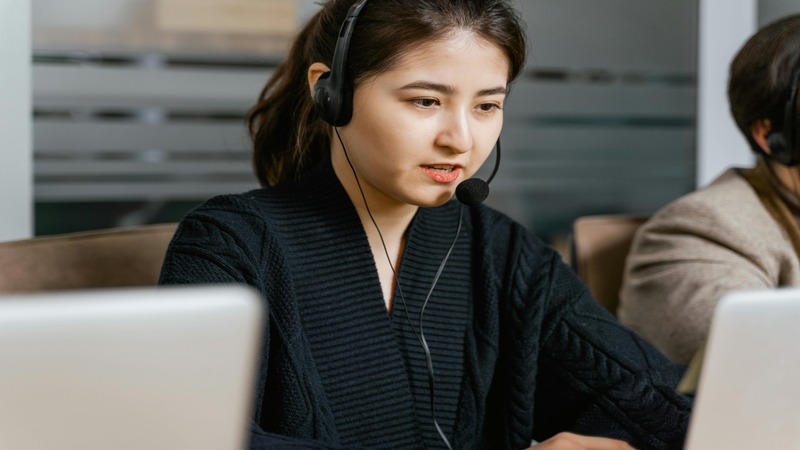 Person in a black sweater and headset sitting in front of a laptop, with another headset-wearing individual partially visible on the right.