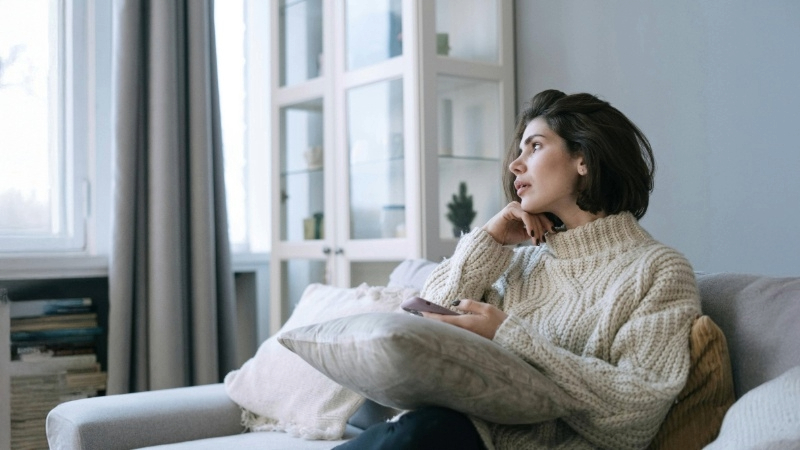 Person in a knitted sweater sitting on a couch, holding a pillow and looking to the side. A glass cabinet, large windows with curtains, and stacked books are in the background.