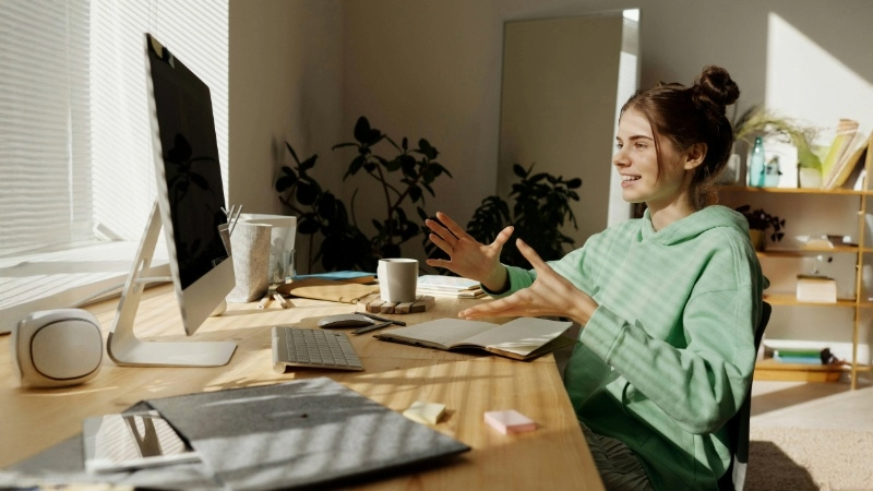 Person sitting at a desk in front of a computer, gesturing with both hands. The desk has a keyboard, mouse, notebook, coffee cup, and stationery. Plants and sunlight are in the background.