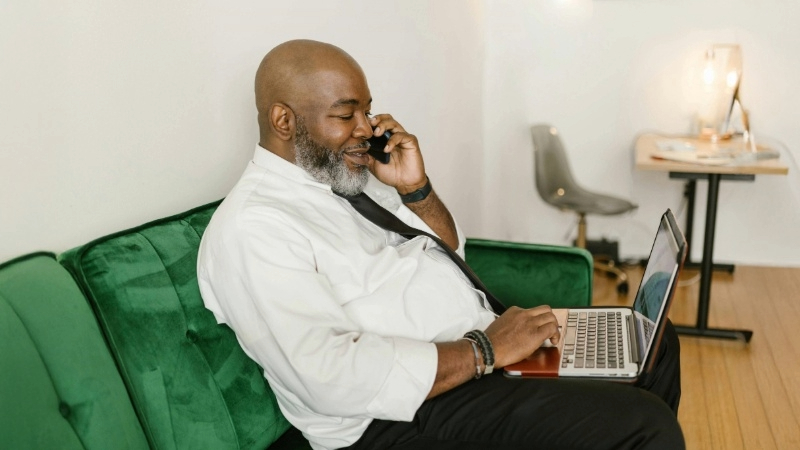 Person on a green couch wearing a white shirt and black tie, using a laptop and talking on the phone. A desk and chair are in the background.