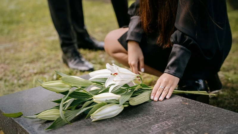 Person kneeling to place a bouquet of white lilies on a gravestone, with another person standing nearby. Scene is set in a cemetery or memorial site.