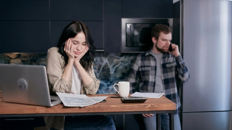 Two people in a kitchen. One is seated at a table with a laptop, papers, and a calculator; the other is standing in the background on the phone.
