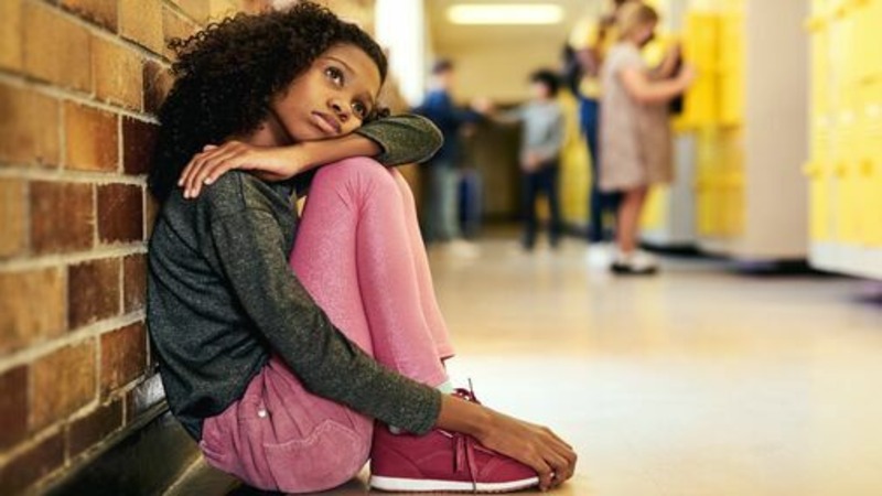 A young girl with long curly hair sits on the ground against a brick wall, hugging her legs and laying her head on her arm, looking up.