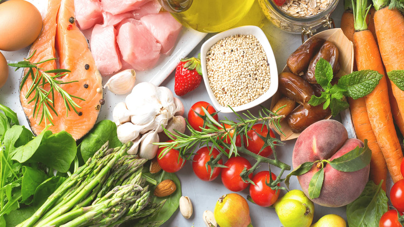 Assortment of fresh foods including salmon, chicken, eggs, vegetables, fruits, nuts, and herbs arranged on a surface.
