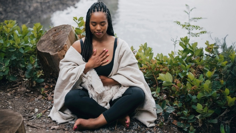 Person with braided hair sitting cross-legged near water, hands in prayer position, in an outdoor setting with plants and a tree stump.