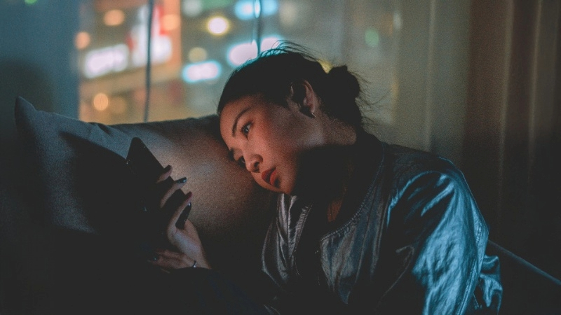 Person sitting on a couch in a dim room, holding a smartphone, with city lights visible through a window.