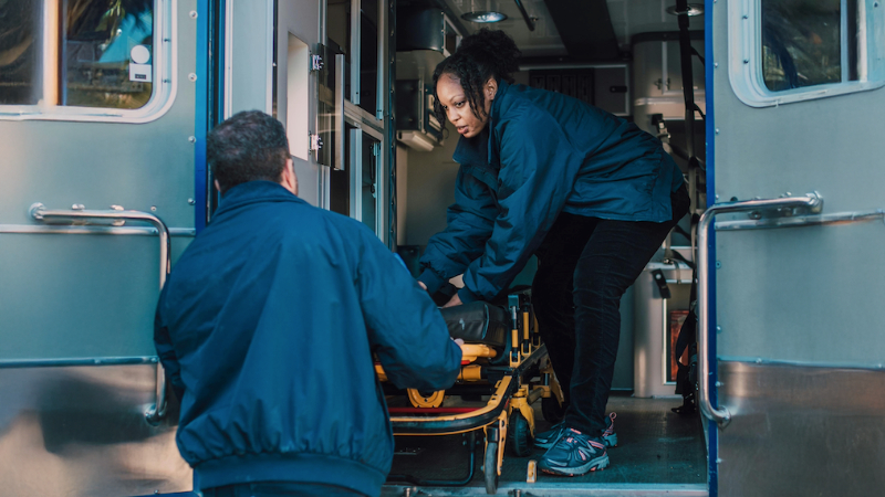 Two people in blue uniforms loading an empty stretcher into an ambulance, with medical equipment visible inside.