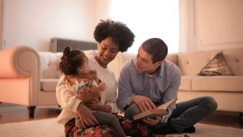 Two parents and a child sitting near a beige couch, reading a large book together.