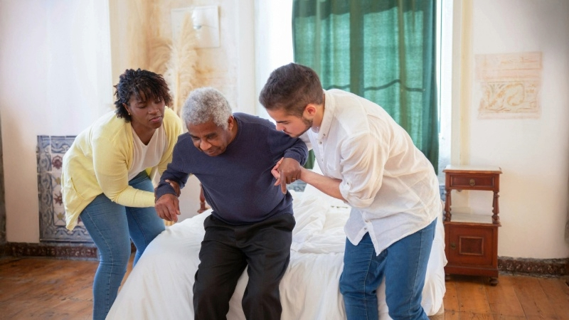 Two people in a room with a white brick wall and a bookshelf; one sits facing the camera with her head in her hands, the other is partially visible facing away.