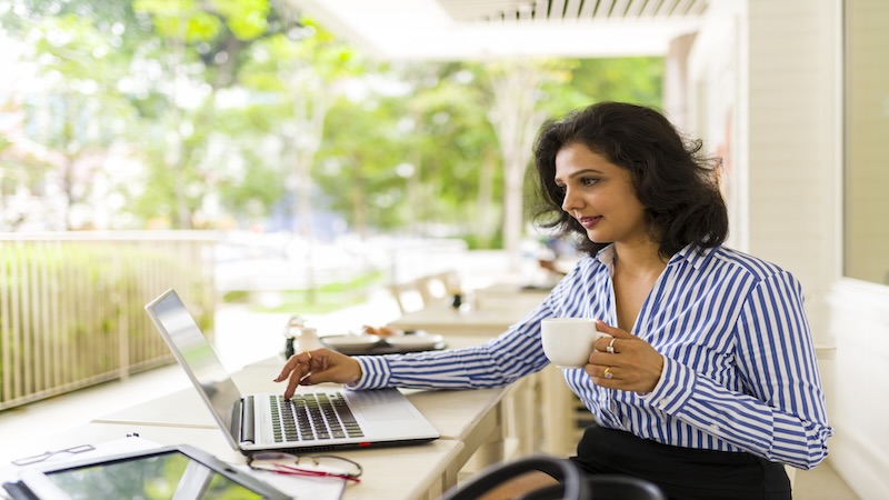 A woman in a striped shirt sits at an outdoor table working on a laptop while holding a coffee cup.